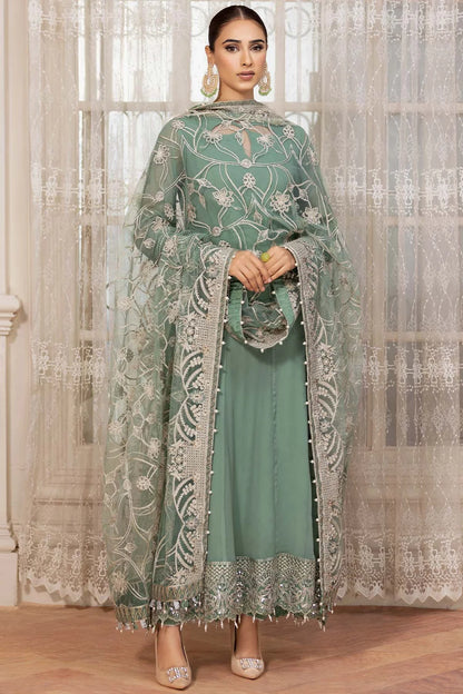 A woman models a traditional three-piece salwar kameez suit in sage green with intricate silver embroidery and a matching sheer dupatta, set against a background of sheer white lace curtains.
