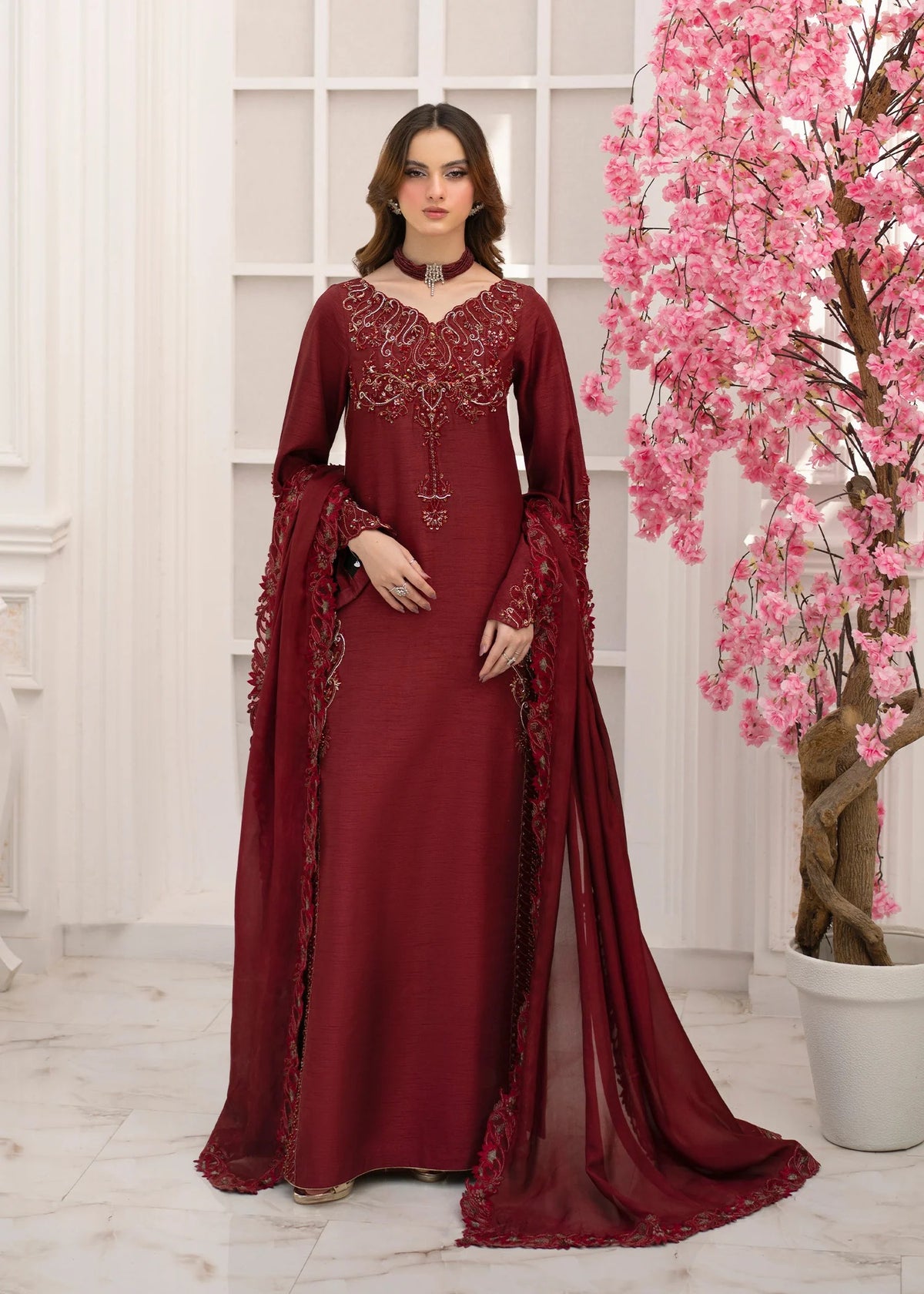 Woman in a burgundy embroidered dress standing next to a pink flowering tree indoors.