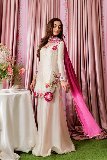 Woman in a white dress with floral embellishments standing in front of pink curtains.