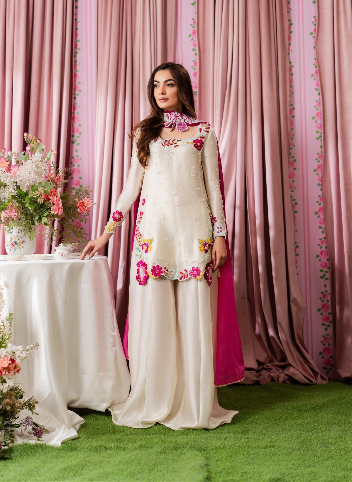 A woman models a traditional South Asian outfit consisting of a cream-colored kurta with colorful floral embroidery and a matching pleated skirt, set against a backdrop of pink floral curtains and green artificial grass.