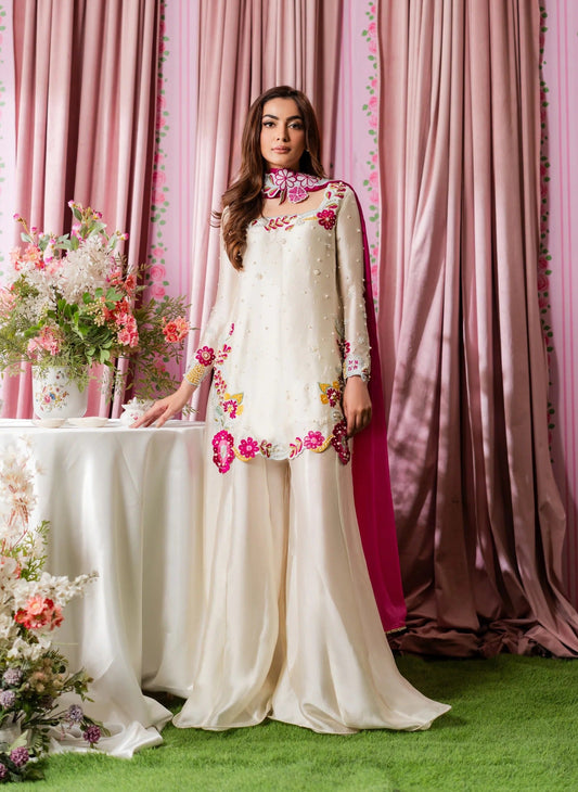 A woman models a traditional white embroidered kurta set with a bright pink dupatta, standing in front of a pink floral draped backdrop.